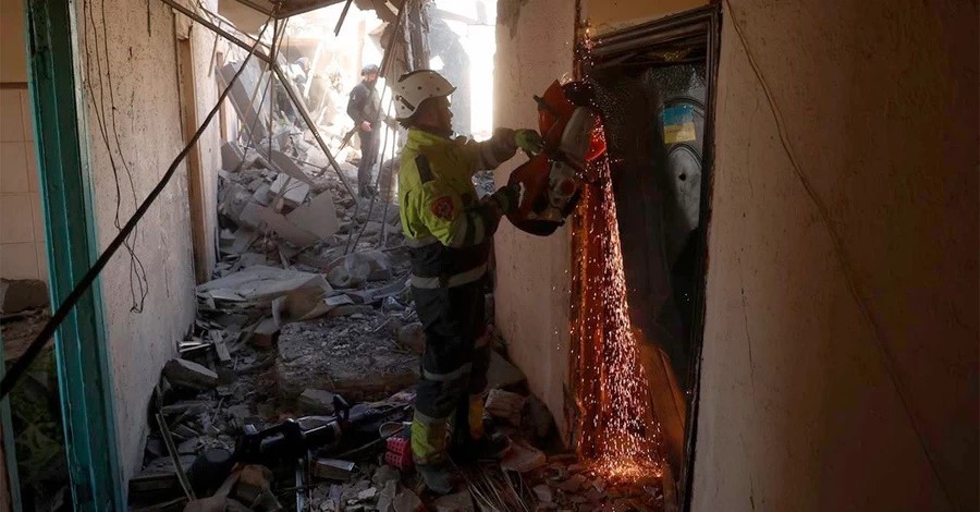 Firefighters in a rubble-filled hallway using a power tool to cut through a metal door as sparks fly, during a rescue or training scenario.